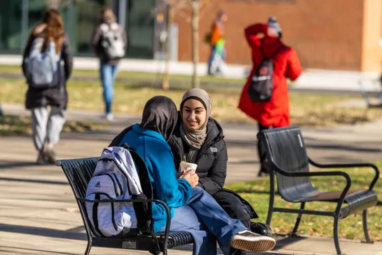 Two people sit on an outdoor bench talking, with others walking by in winter clothing.